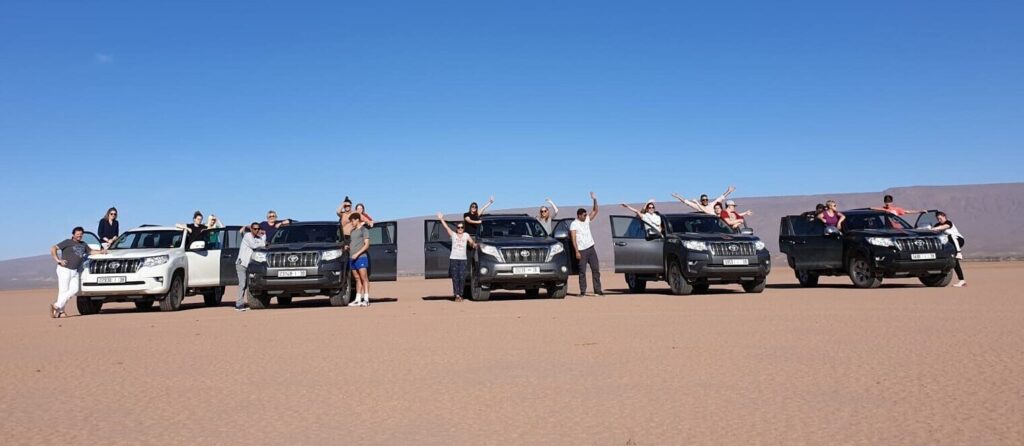 DesertChigagaTours - Gallery - Five 4x4 cars parked next to each other in the Sahara. Four to five people around each car waiving for a group photo