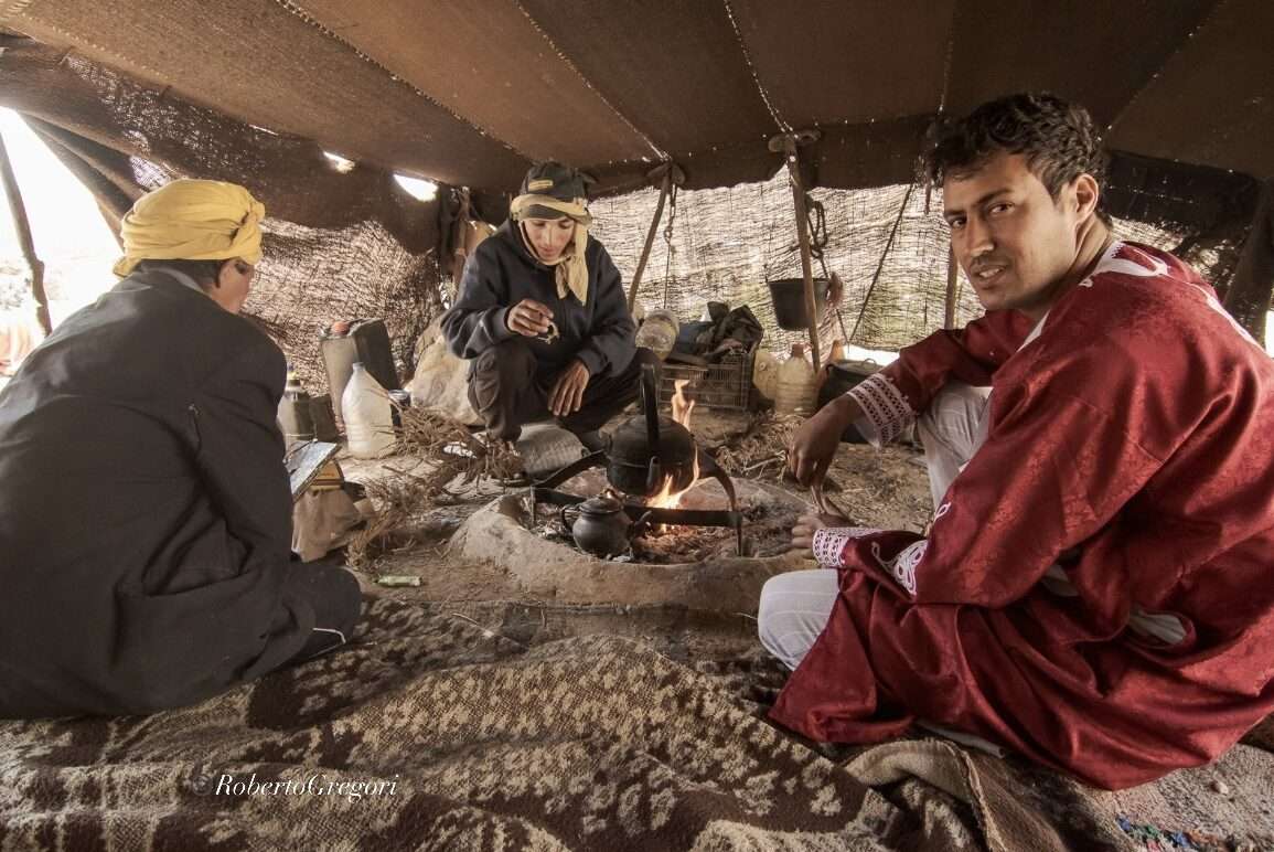 3 guys under a nomad tent in the maroccan mountains. One has a red Djellaba. they are preparing tea on the fire