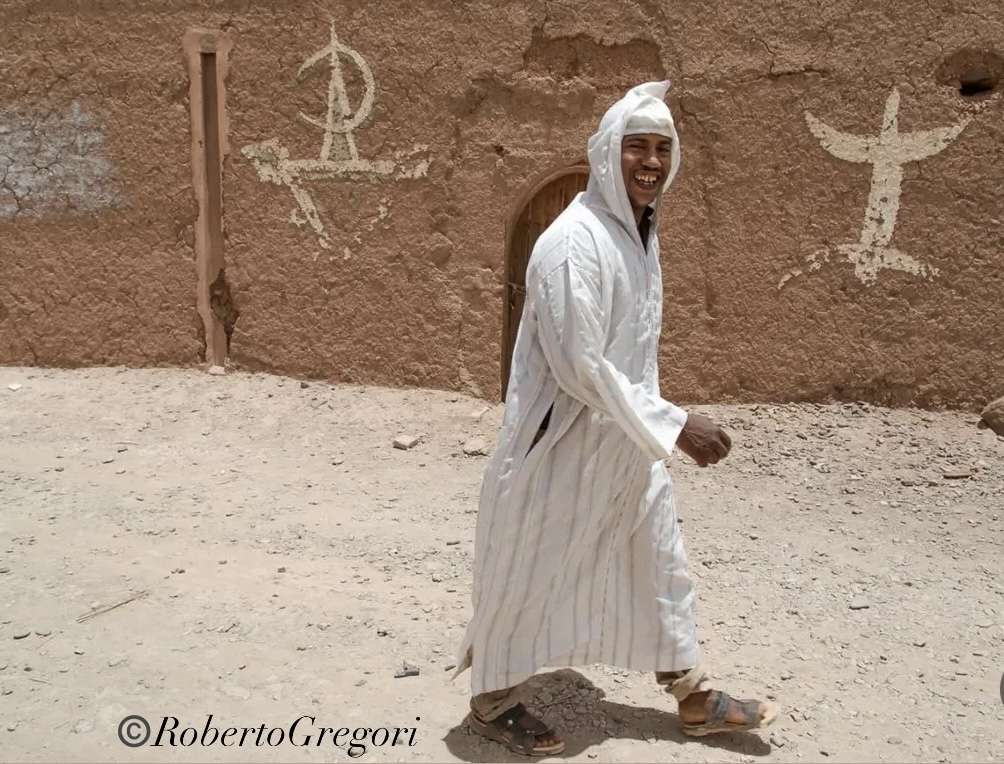a berber man wearing a white djellaba walks in front of a mud house smiling. Two tyical berber white signs on the walls of the house