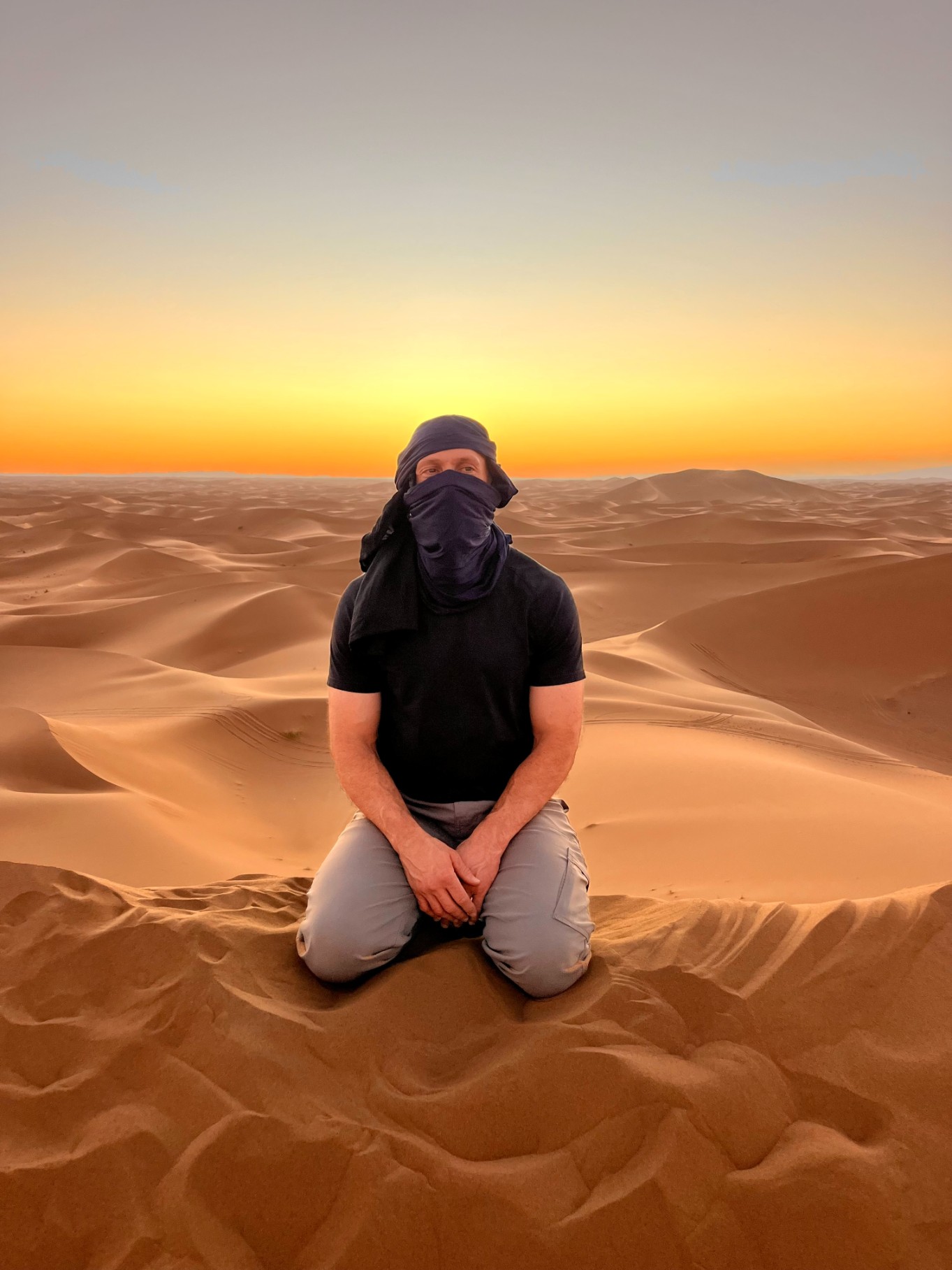 A man with blue turban sitting on top of a sad dune at sunset. Orange sunset colors on the back.