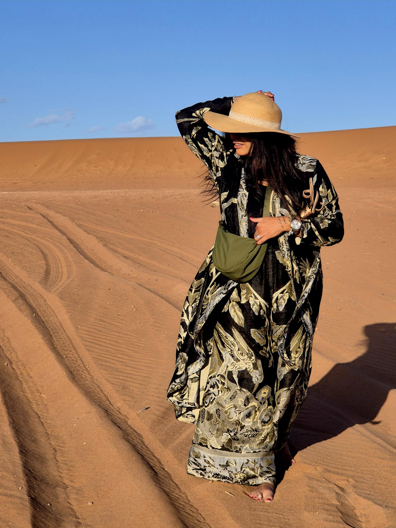 A young woman walking in the desert with blak and olive kaftan, olive purse and straw hat. Orange-red sand dunes on the back