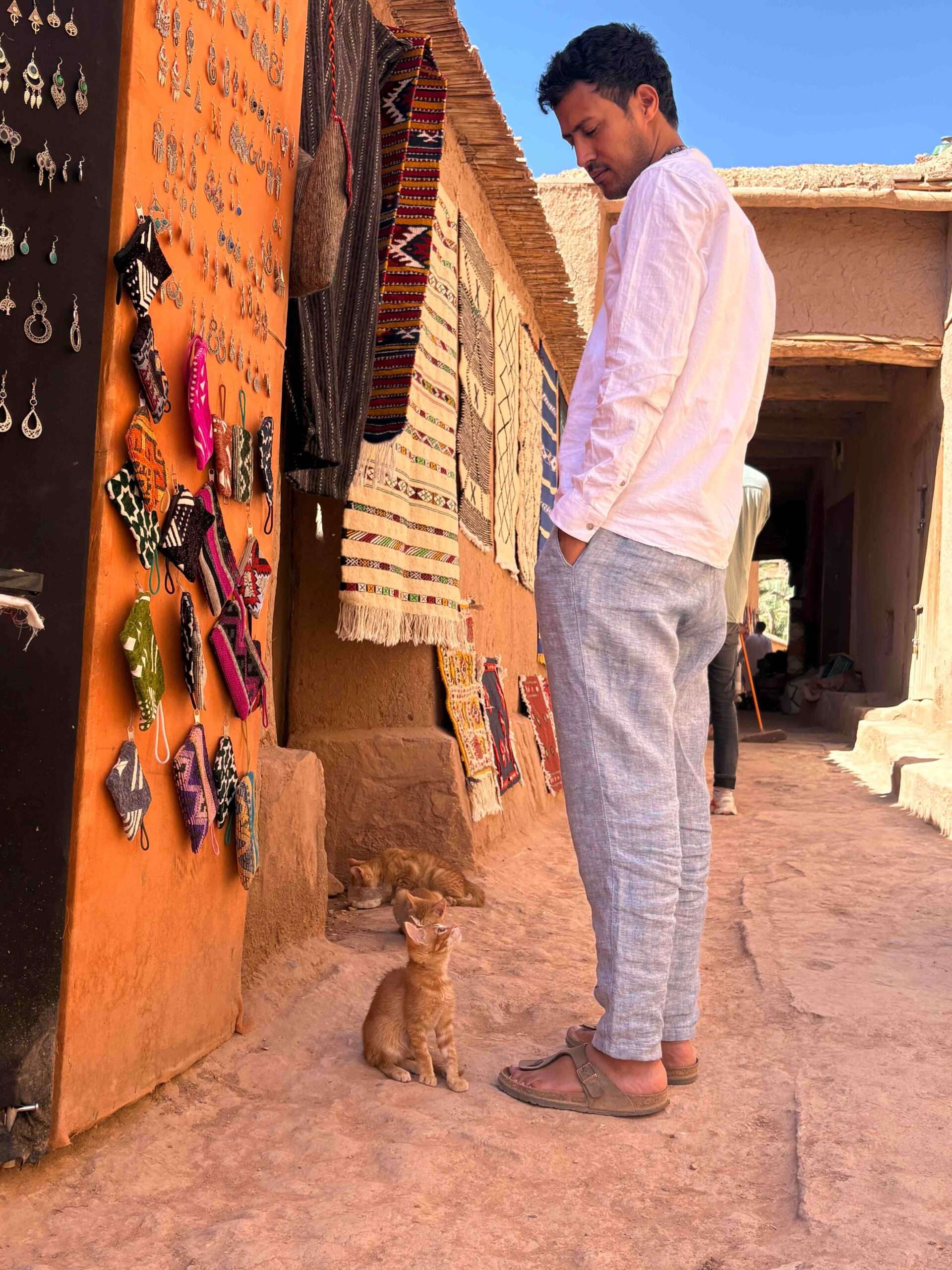 Young Moroccan man, on the street of Aït Benhaddou, in white shirt and light blue cotton pants standing and watching an orange tiny cat close to his feet. The cat is watching at him.