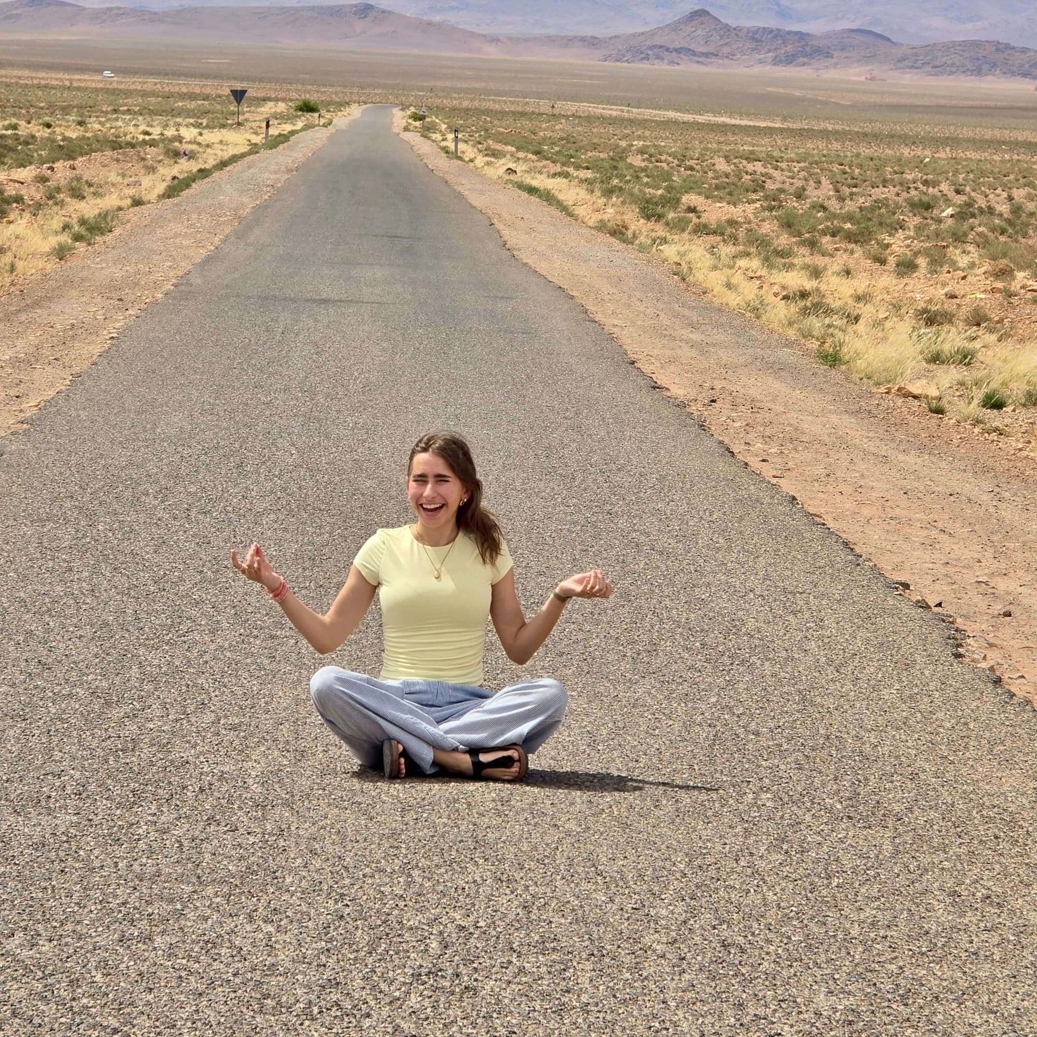 yung woman in yellow T-shirt and light blue cotton pants, long brown hair, smiling, sitting in the middle of a straight road in the desert.