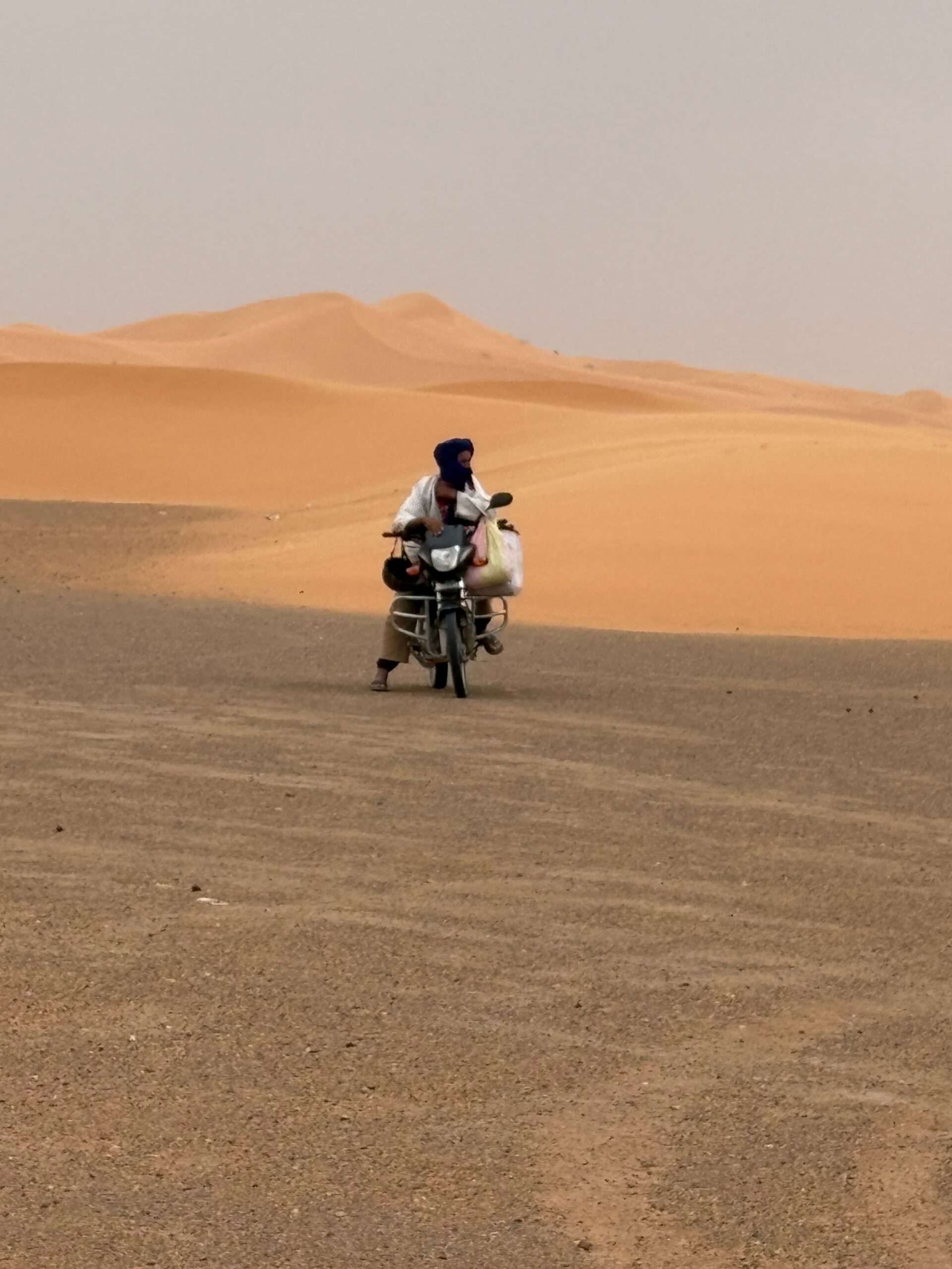 Touareg in Merzouga with blue turban and white djellaba, on a motorbike watching the dunes at sunset